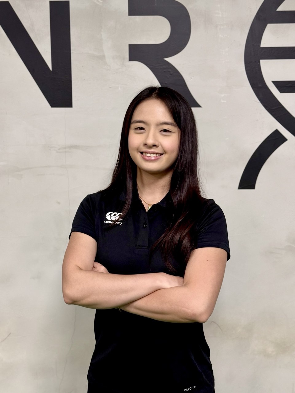 A female trainer with long dark hair stands with arms crossed, wearing a black Canterbury polo shirt in front of a light gray wall with large black lettering.